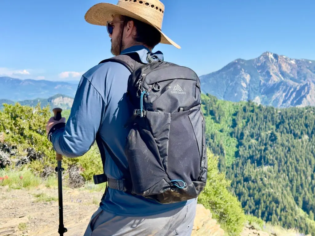A man stands on a trail in front of mountains carrying a black Gregory Kiro daypack.