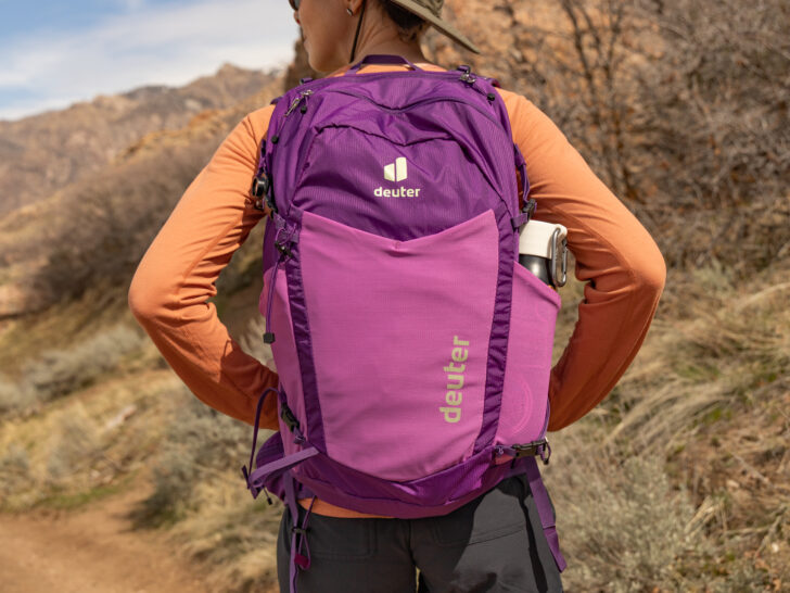A purple Deuter daypack on a woman's back as she stands on a sunny trail.