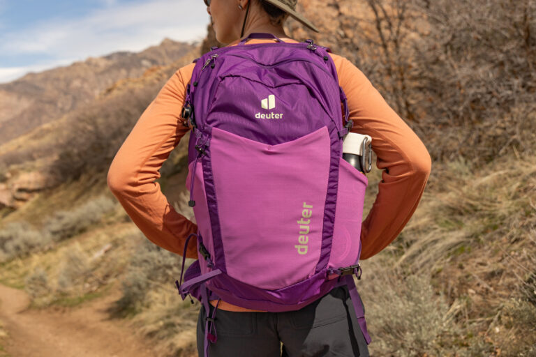 A purple Deuter daypack on a woman's back as she stands on a sunny trail.