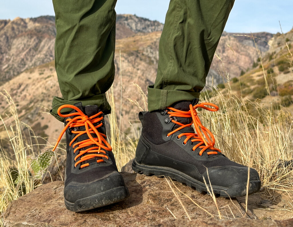 Black Bahé Boots with orange laces. On a hiker's feet as she stands on a rock before mountains.