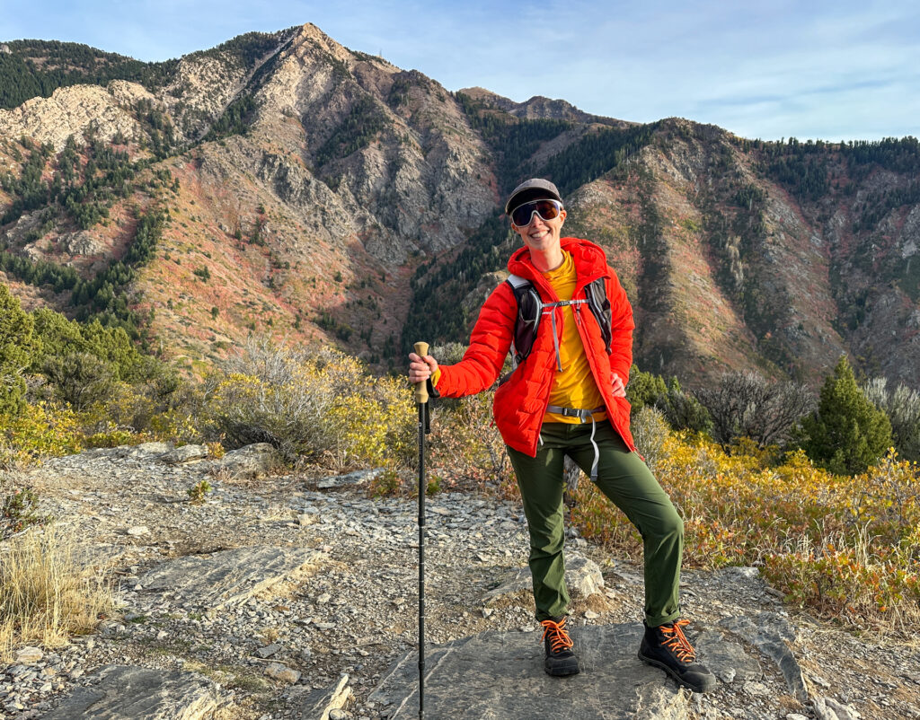 A woman with a backpack, jacket, and Bahé boots standing in front of a peak.