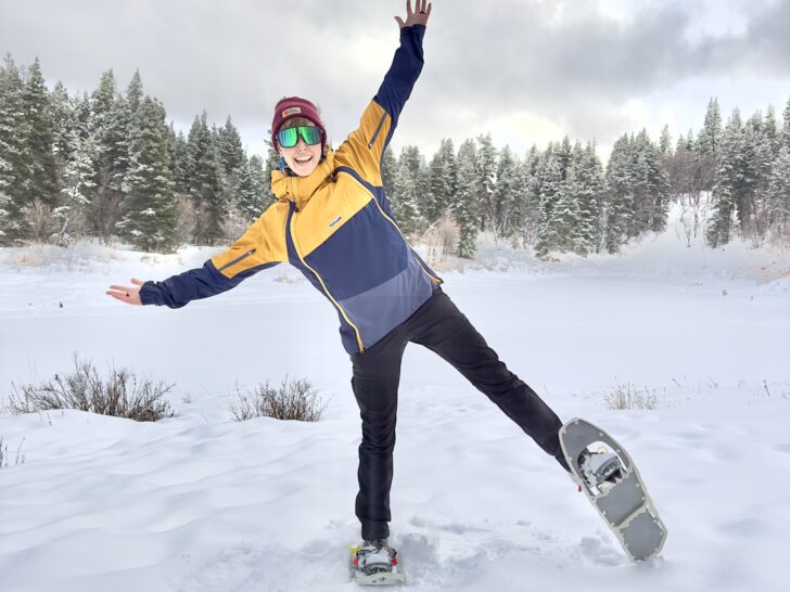 A woman poses in snowshoes in front of a snowy lake and pine trees.