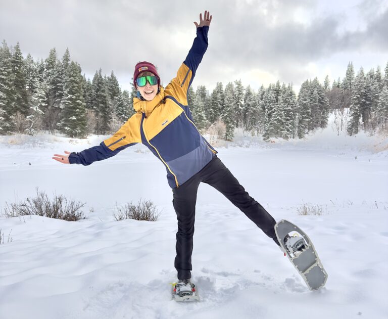 A woman poses in snowshoes in front of a snowy lake and pine trees.