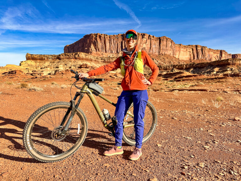 A woman stands in front of a mesa with her bike on White Rim Road in Canyonlands.