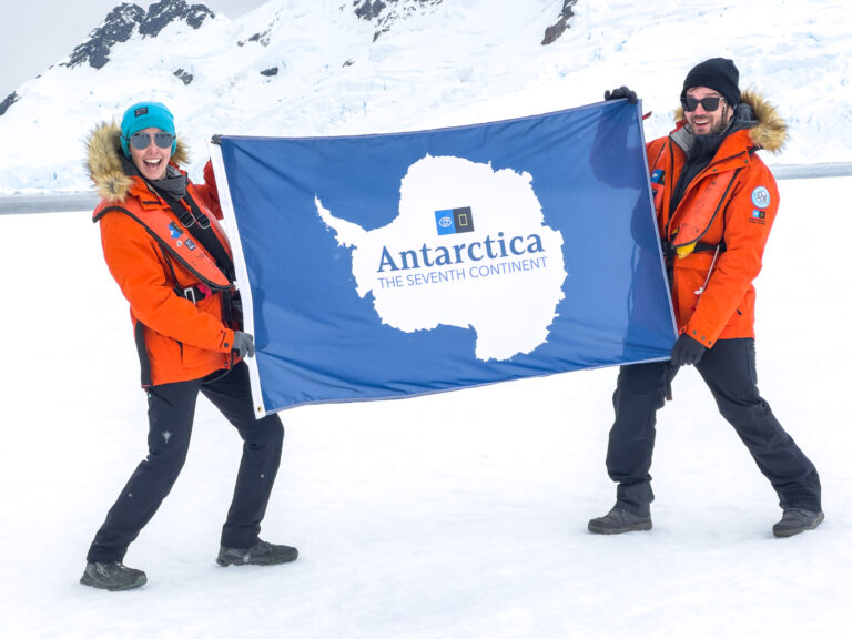 A man and woman in parkas smile and hold an Antarctica flag while standing on sea ice.