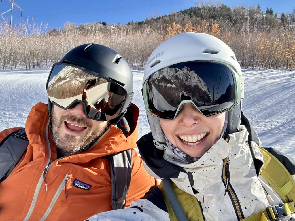 A man and woman smile in snowboard helmets and goggles.