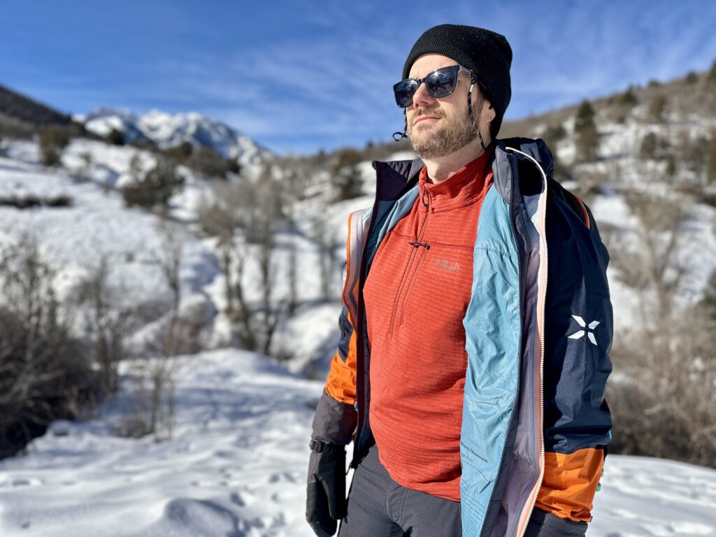 A man in layers and a Rab Ascender Pull-On stands in front of snowy mountains.