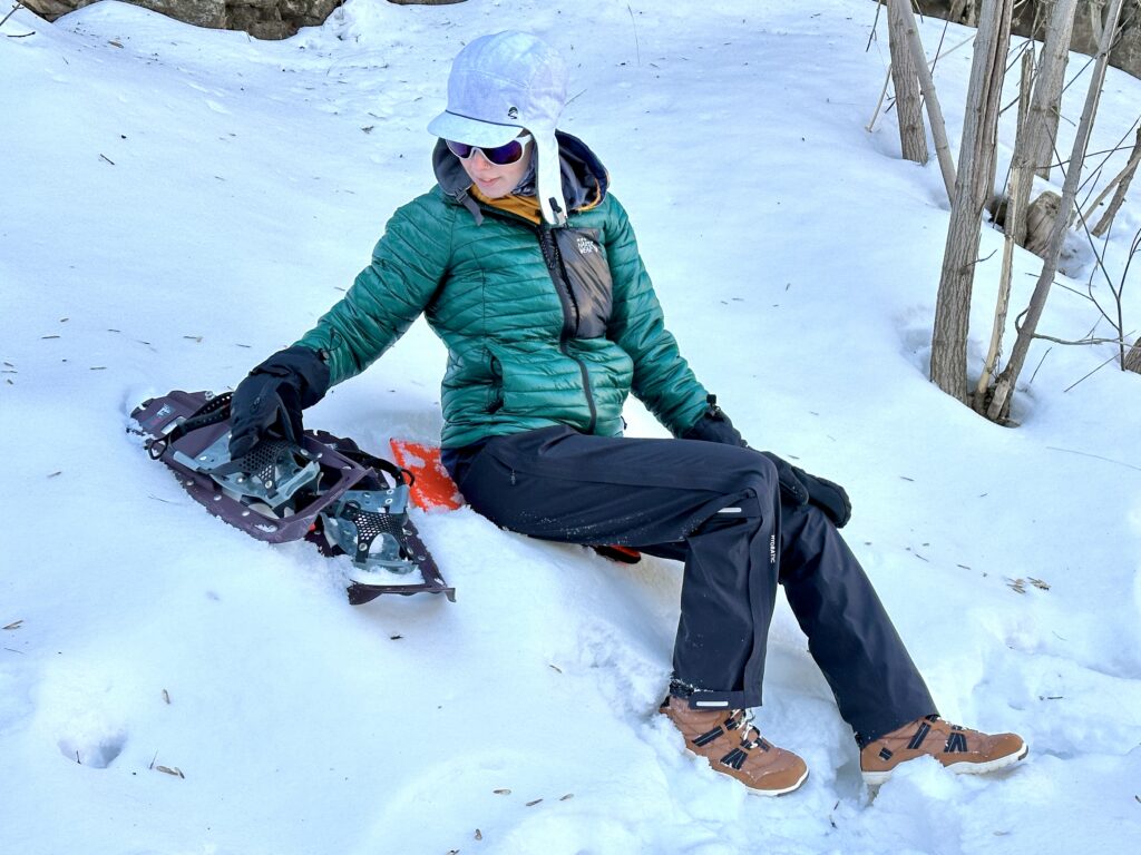 A woman sits in the snow next to snowshoes in Fjallraven waterproof Pants.