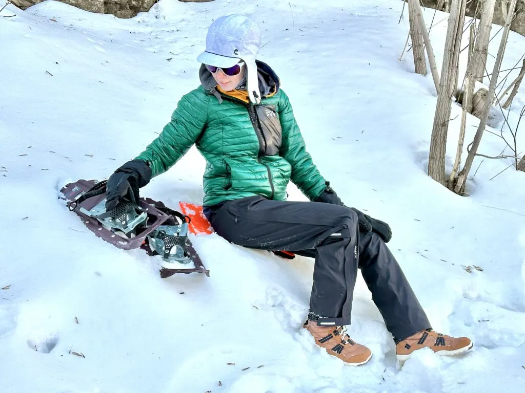 A woman sits in the snow with snowshoes. She is wearing Fjallraven High Coast Hydratic Trousers.