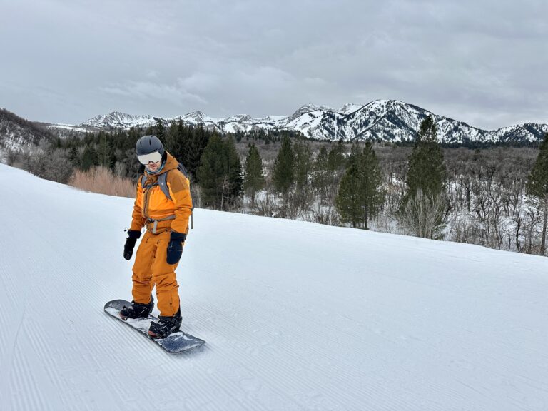 A man in a Picture Organic Clothing ski jacket and bibs snowboards with mountains behind him.