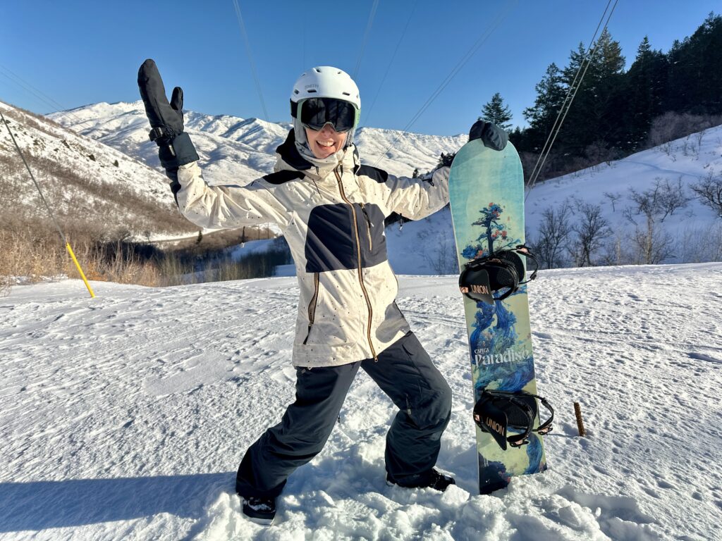 A woman poses with a snowboard in front of mountains.