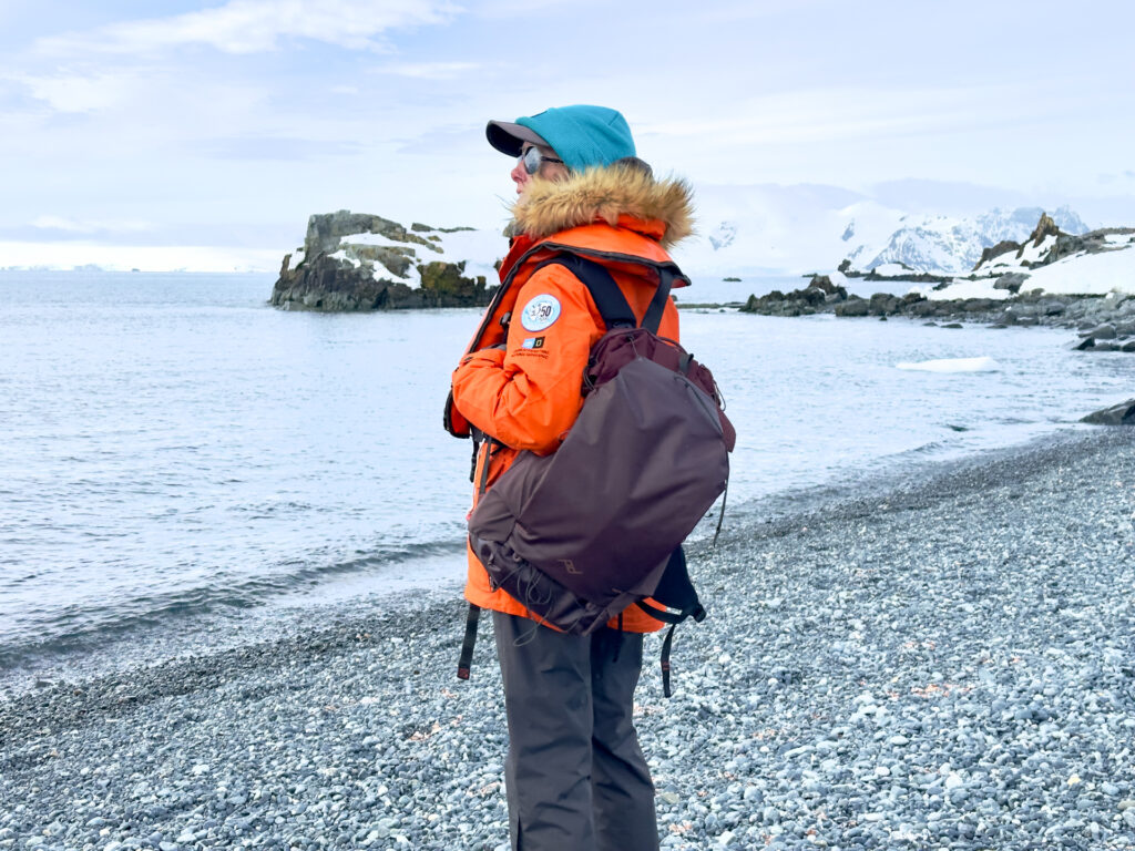 A woman in a park stands on a rocky beach in Antarctica with a Peak Designs Outdoor 25L Backpack.