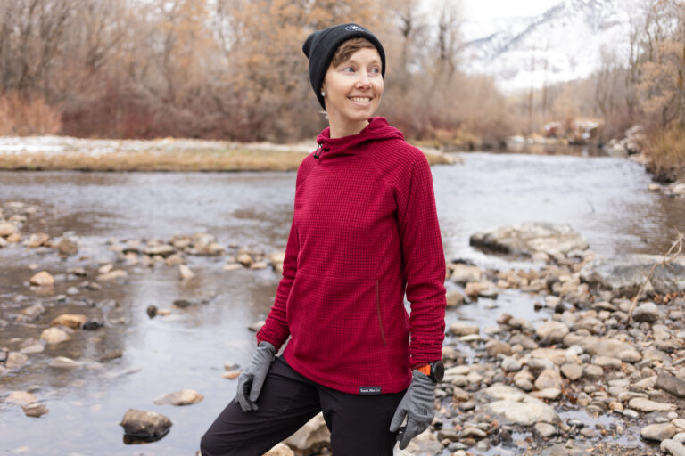 A woman smiles in front of a river. She wears a Squak Microgrid fleece hoodie.