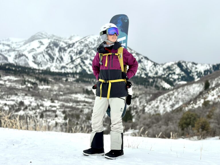 A woman in a jacket and bibs stands in front of mountain with a backpack and snowboard on her back.