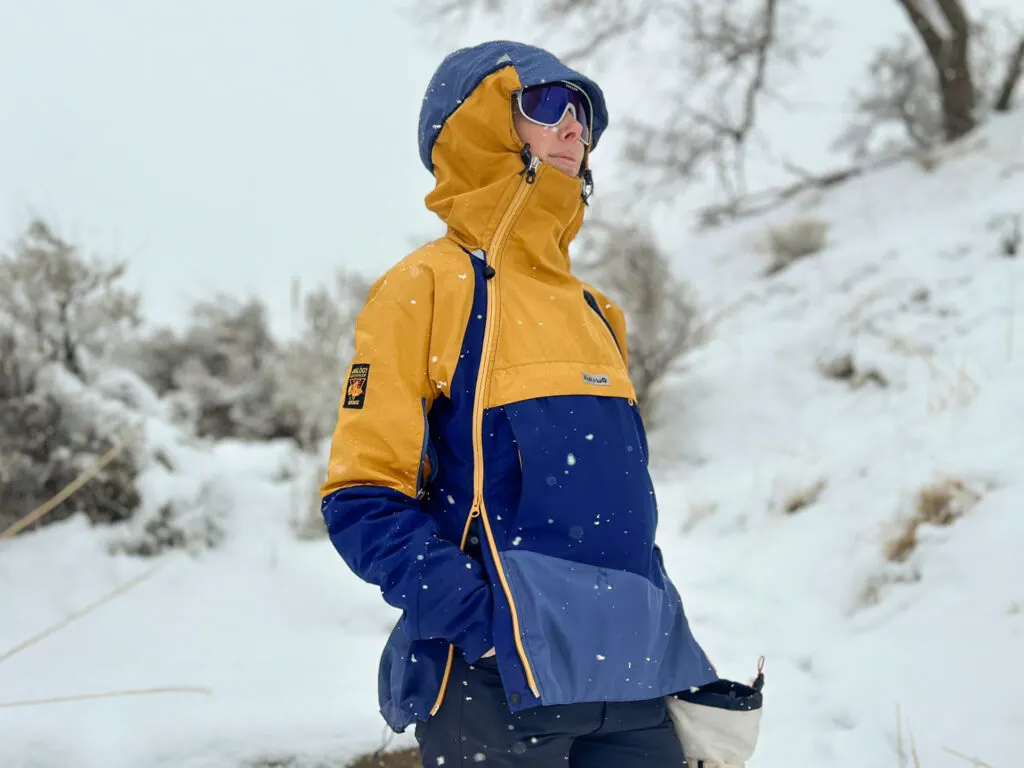 A woman wears a Paramo Velez Evolution Hybrid Smock in the snow. The side zippers are unzipped from the bottom up and her hands are in her pants pockets.