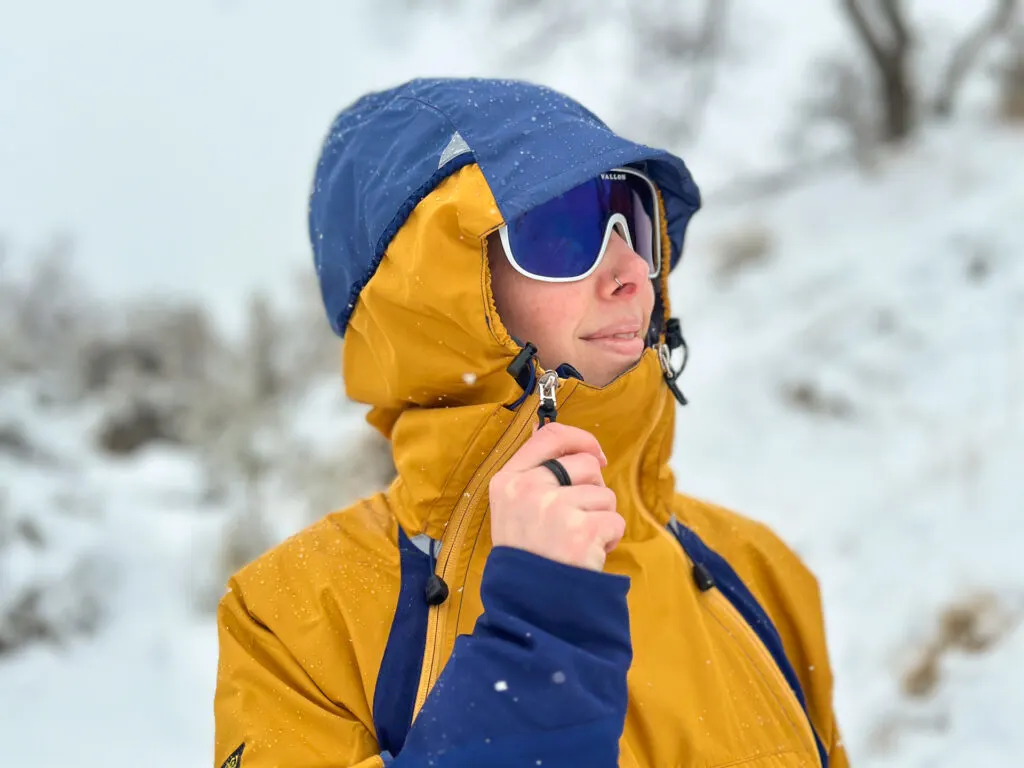 A woman wears a Paramo Velez Evolution Hybrid Smock in the snow. The side zippers are fully zipped and the hood is cinched and fitted.