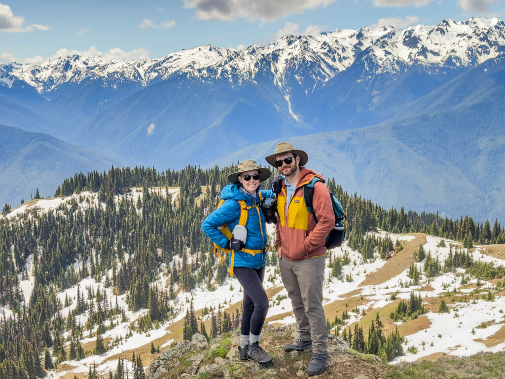 A couple in backpacks stand in front of mountains at Olympic Peninsula National Park.