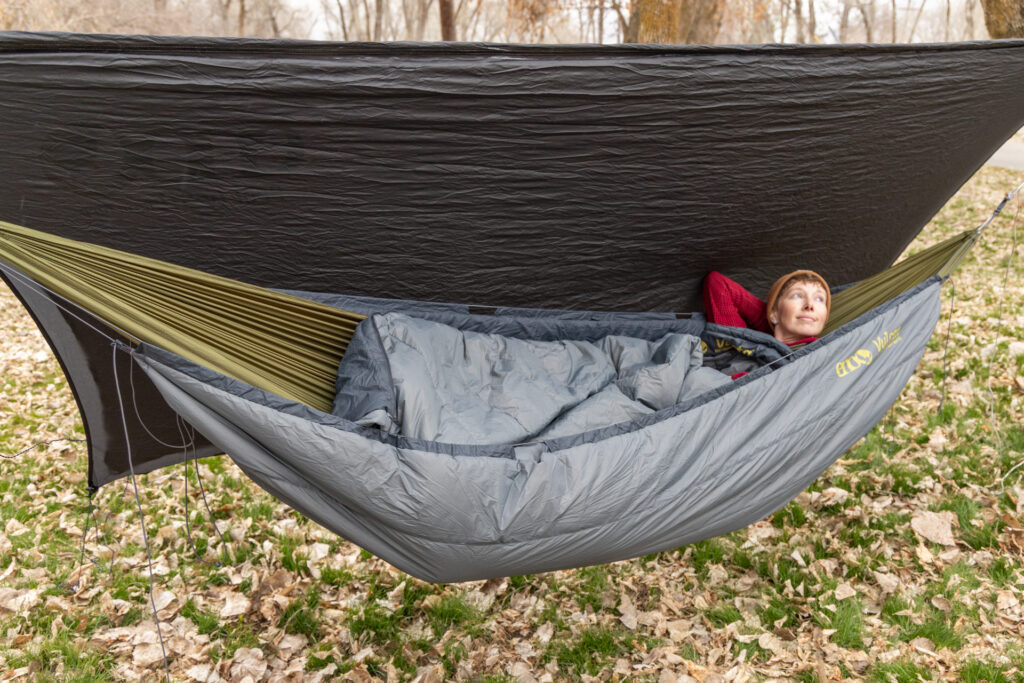 A woman lays in The Eno SubLink Hammock Camping System, complete with underquilt, topquilt, and tarp flipped to one side.