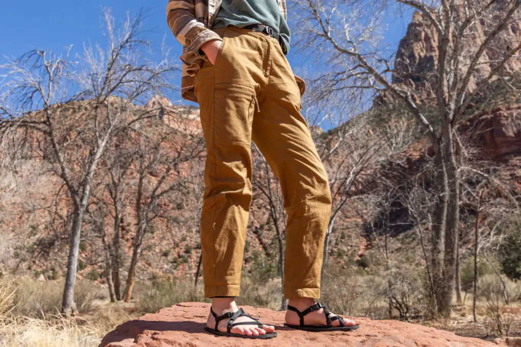 A woman's lower half wearing the Mammut Tamaro Pants as she stands on a rock in front of cliffs.