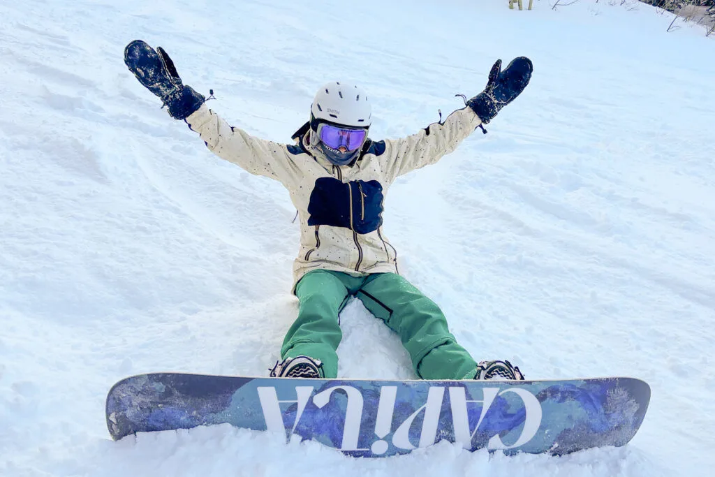 A woman sings in the snow with her snowboard on her feet. She is wearing the Gnara x Burton bibs.