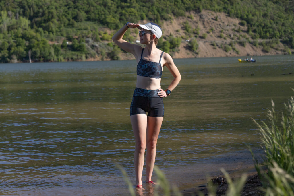 A woman stands with a paddle board in front of a lake wearing a Finisterre Sula bikini top and Aurora swim shorts.