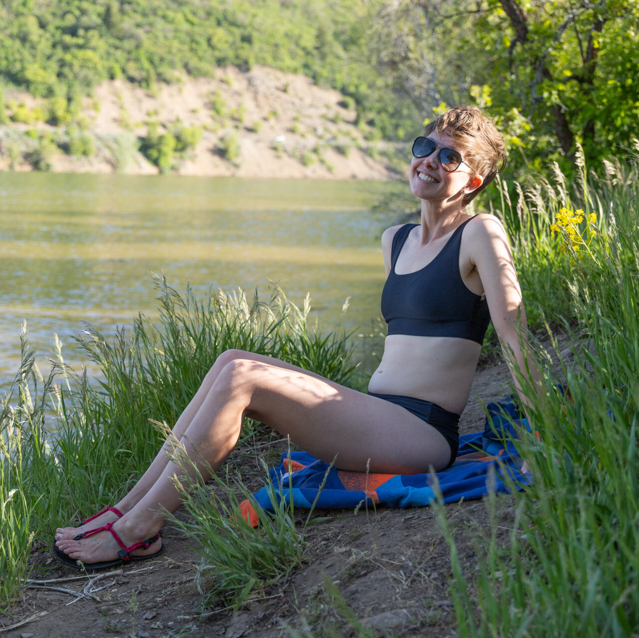 A woman sits by a lake wearing a Free Fly Apparel Scoop Bikini sustainable swimsuit.