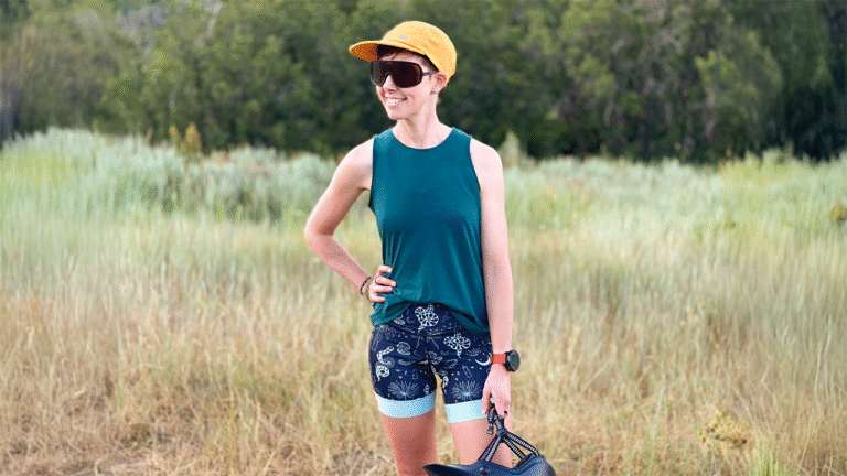 A woman stands in a field wearing MTB clothing, a cap and Obraz sunglasses.