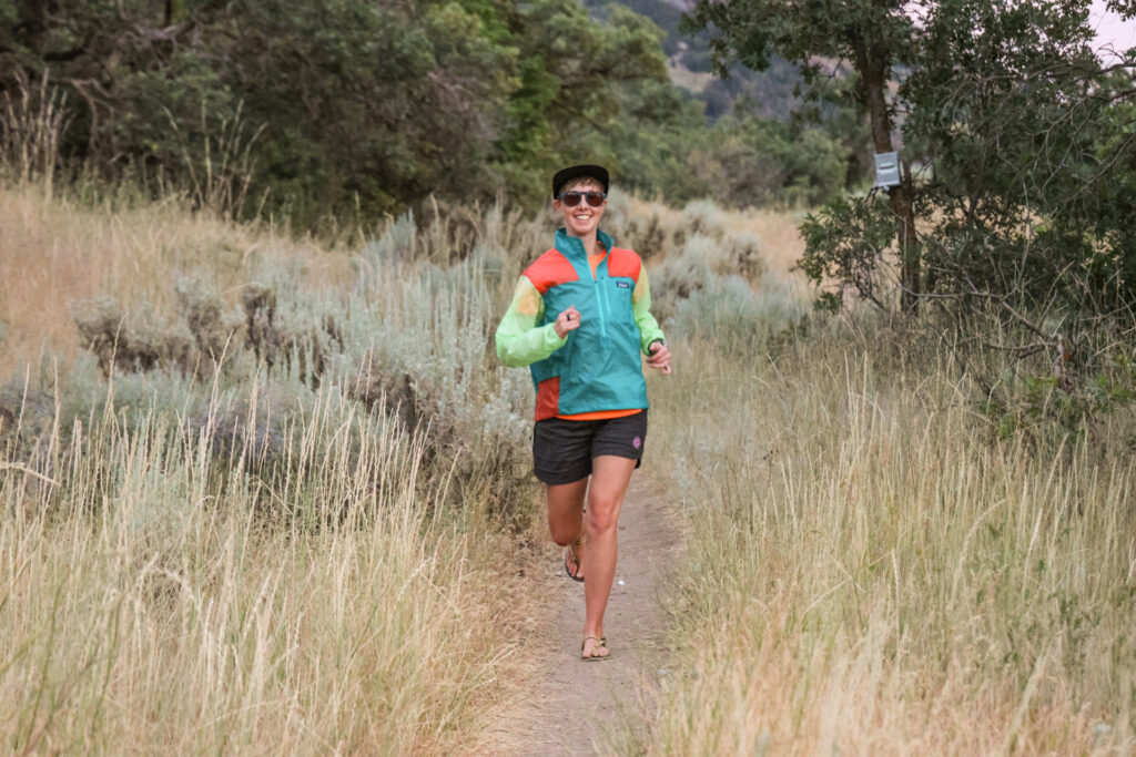 A woman trail runs in a windbreaker and barefoot running sandals.