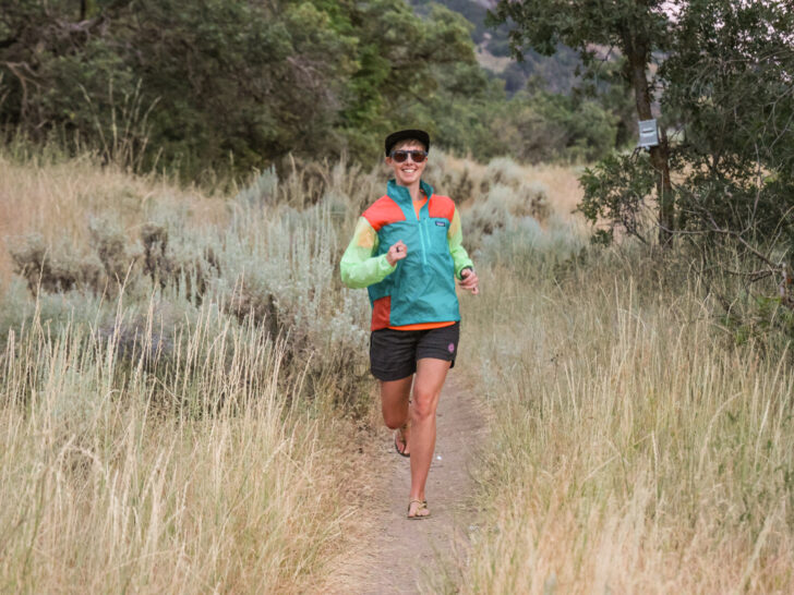 A woman trail runs in a windbreaker and barefoot running sandals.