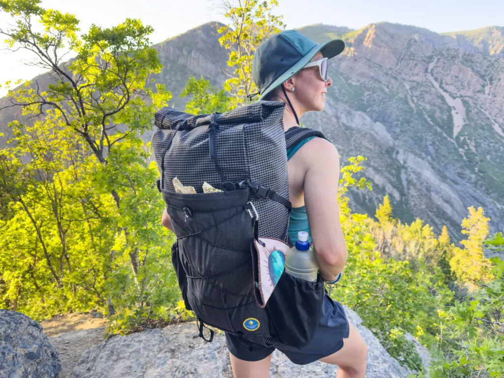 A woman stands on a hiking trail wearing the Symbiosis Gear Aspen backpack.