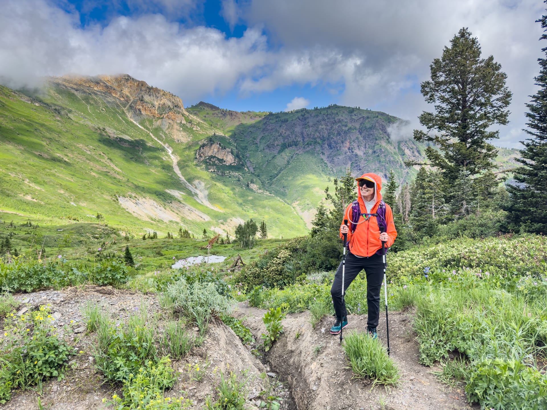 A woman in a windbreaker and backpack stands on a hiking trail with mountains behind her.