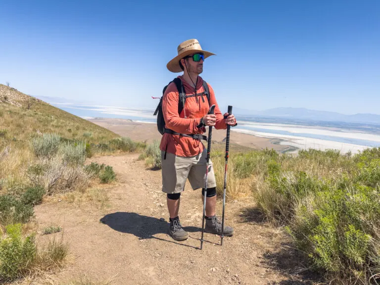 A man wears a backpack and stands on a trail with trekking poles.