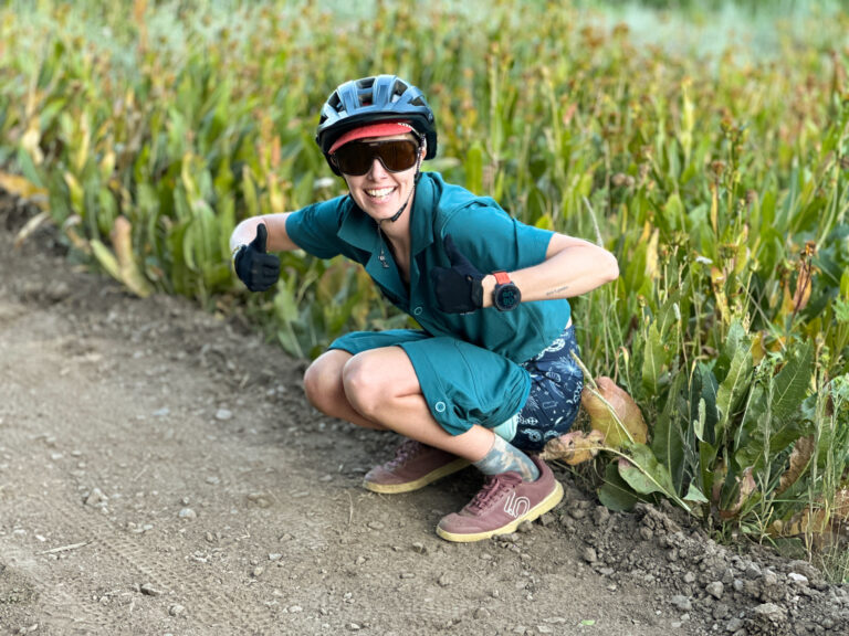 A woman in Shredly mountain bike apparel, namely the Drop Tail Trail Romper, squats on the side of a trail.