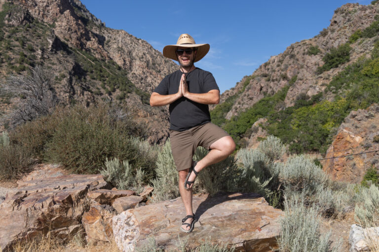 A man in a hat does a standing yoga pose on a rock in front of mountains. He is wearing Astral hemp clothing.