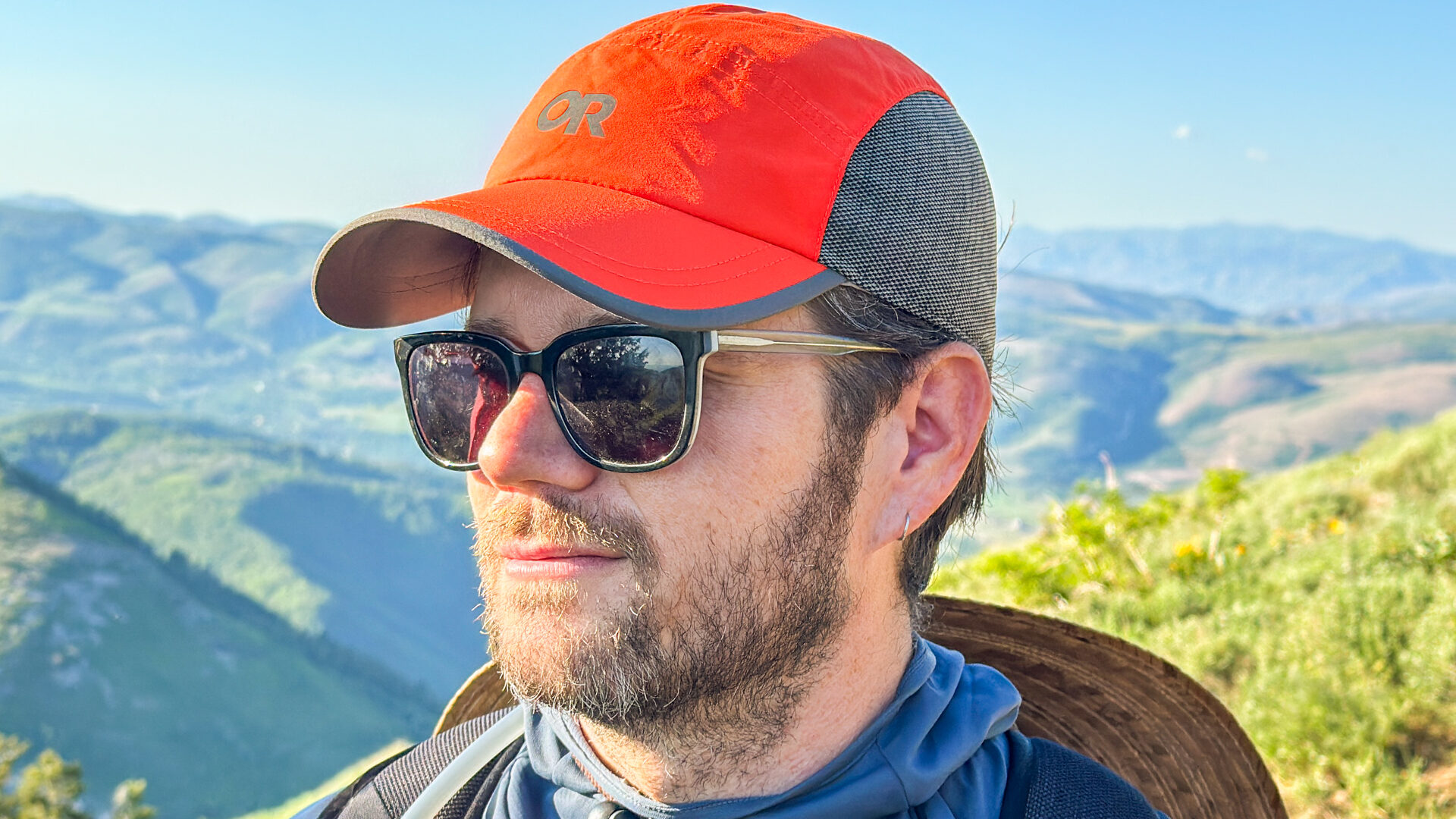 A man wearing sunglasses on a mountain wearing an orange and grey Outdoor Research Swift Cap.