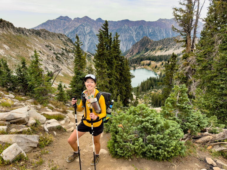 A woman with a backpack stands high above an alpine lake with mountains in the distance.