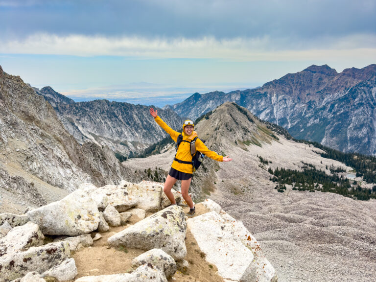 A woman does a silly pose on a peak with mountains in the background.