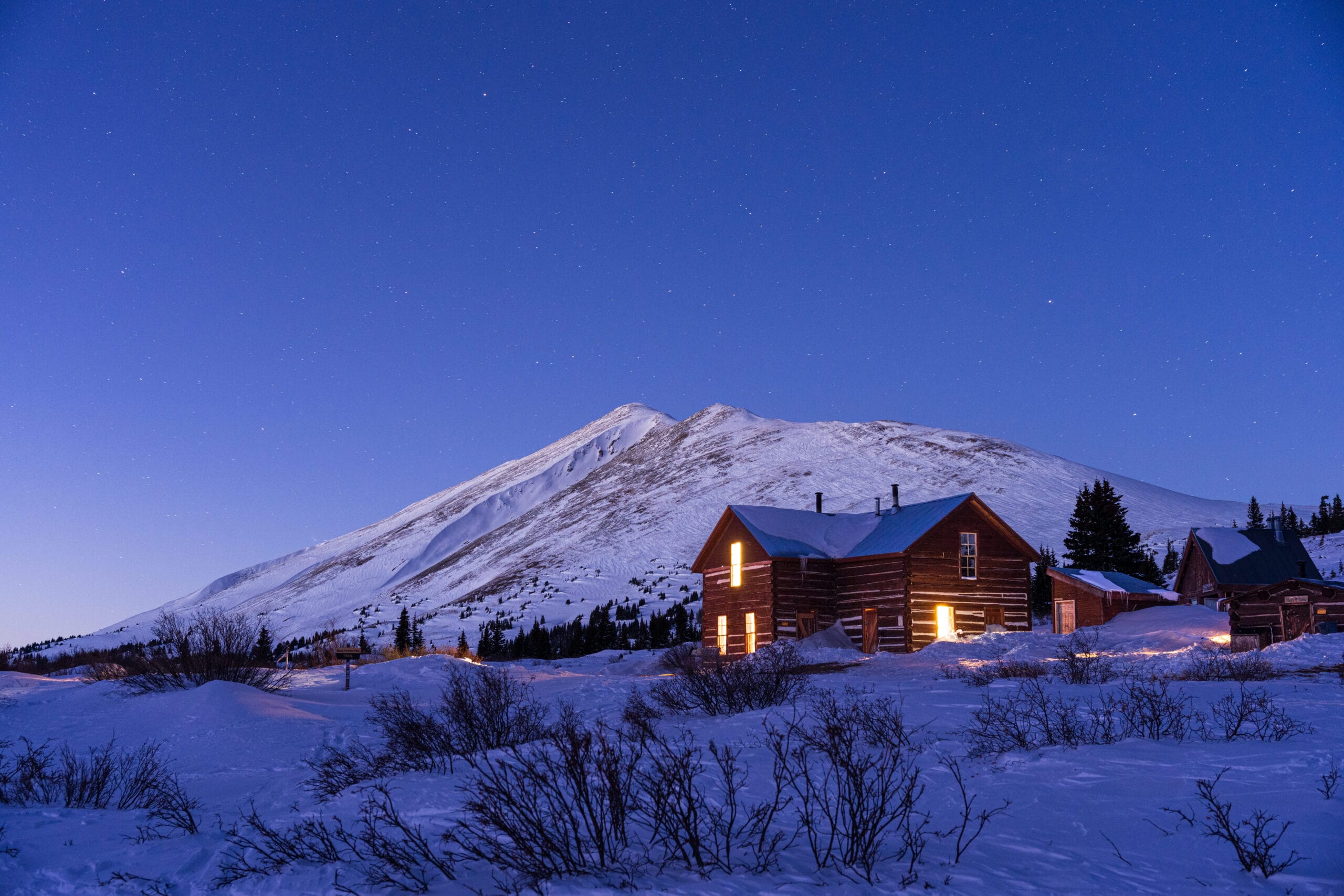 A cabin is lit up in the dark with a snowy mountain behind it.