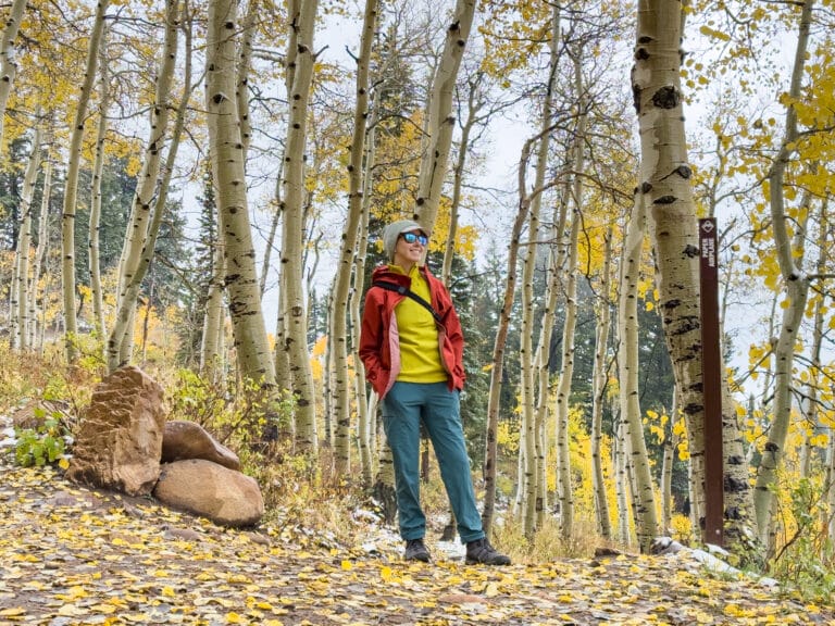 A woman in several layers and a rain jacket stands on a trail surrounded by fall colors and aspen trees.