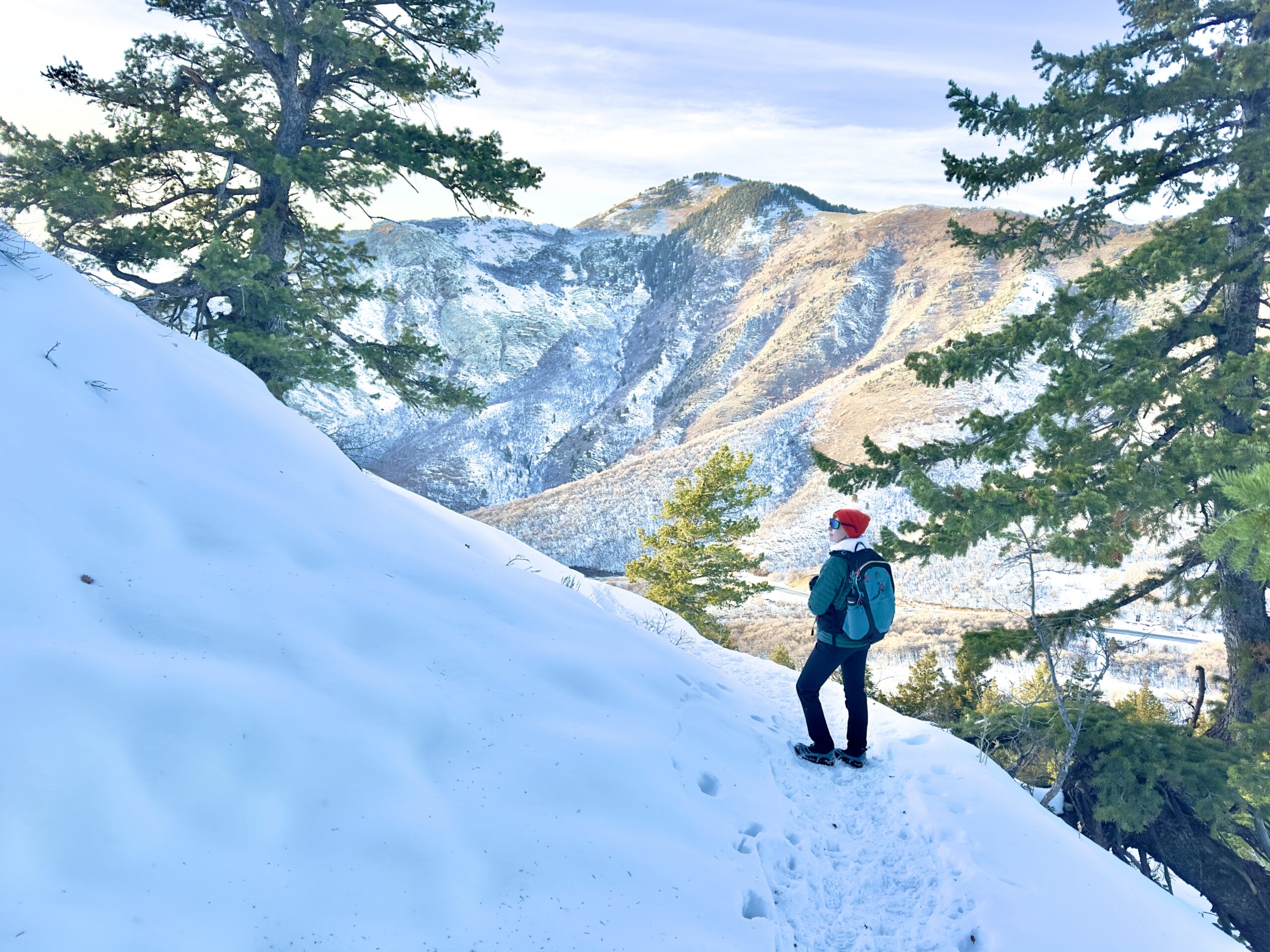 A woman in winter layers stands on a snowy trail with mountains in the background.