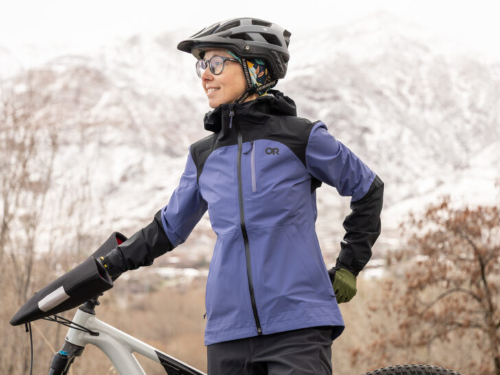 A woman in a helmet wears an Outdoor Research Freewheel Stretch Jacket next to a bike.