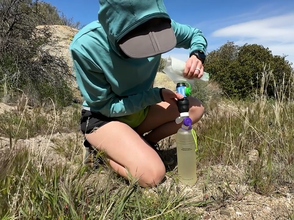 A woman bends over a Sawyer water filtration system in the backcountry.