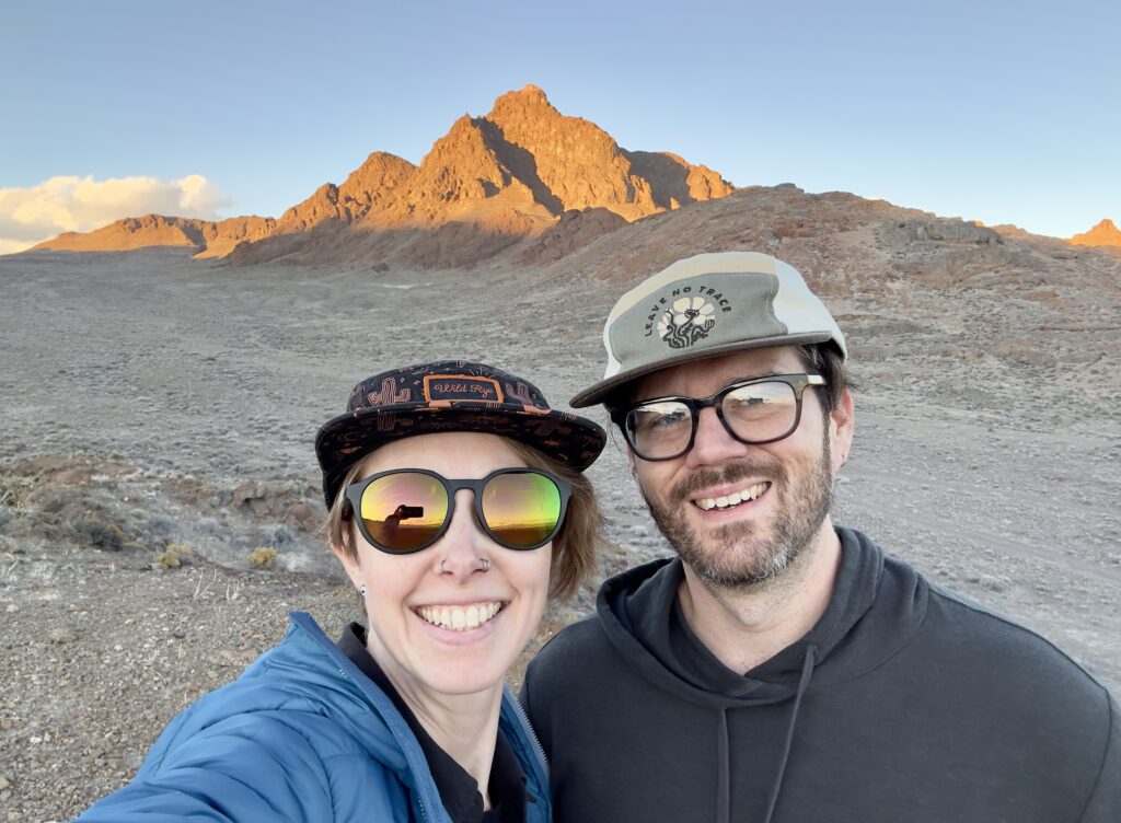 A man and woman smile in front of a peak. One is wearing prescription glasses, the other prescription sunglasses.