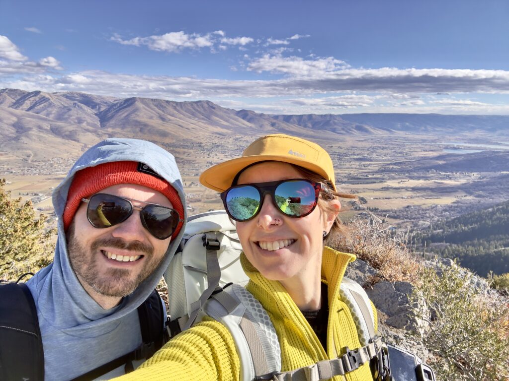 A man and woman smile in front of a peak. One is wearing sunglasses, the other prescription sunglasses.