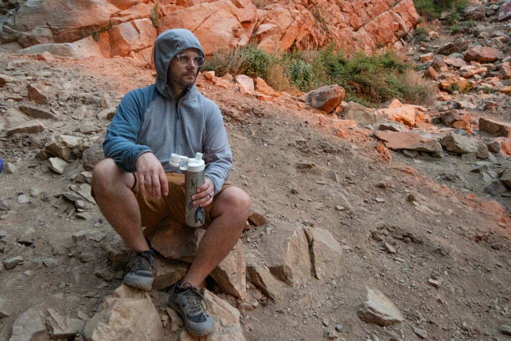 A man in a hoodie sits on a rock on a hiking trail wearing wide toe box hiking shoes from Merrell.