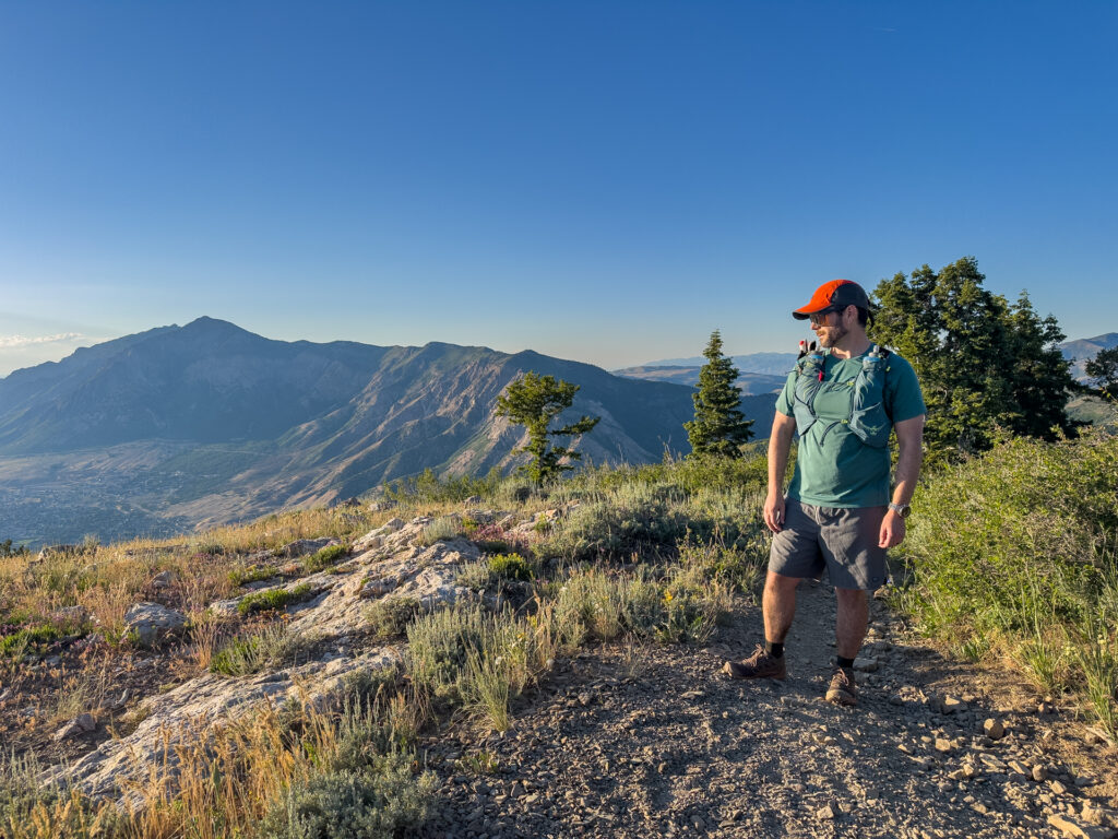A man stands on a mountain peak with peaks in the background. he is wearing running clothing and a trail hydration vest.