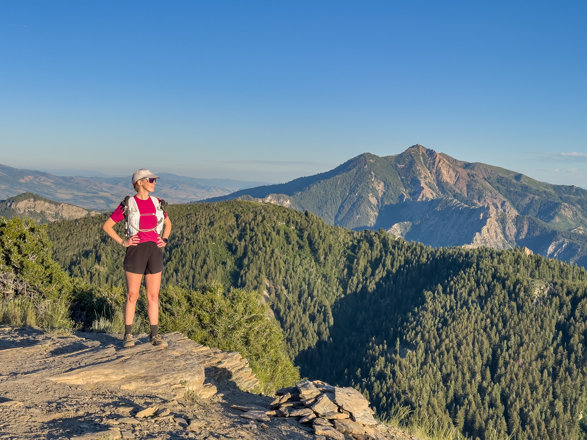 A woman stands on a mountain peak with mountains in the background. She is wearing trail running vest.