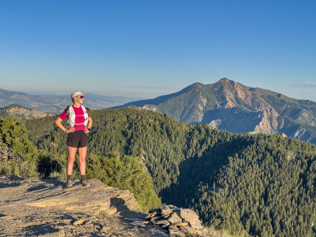A woman stands on a mountain in hiking clothing and barefoot shoes with a wide toe box.