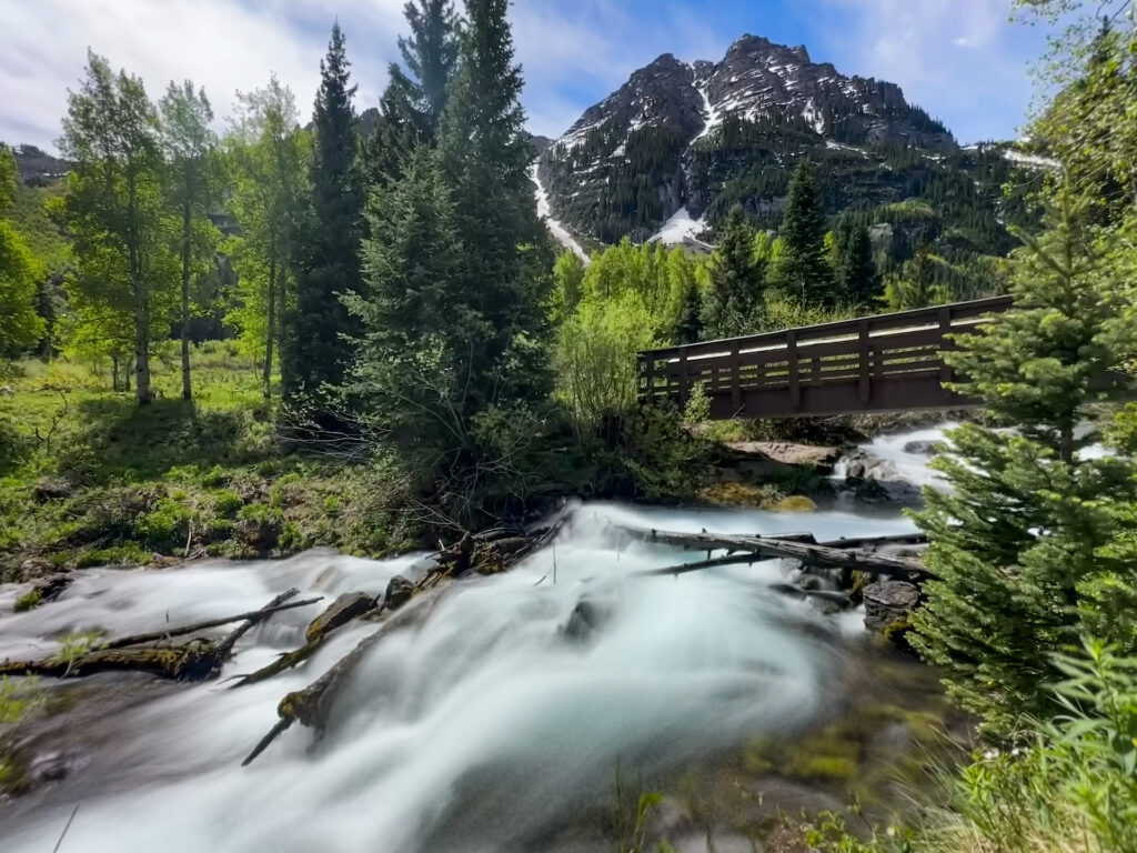 Maroon Bells Hike, Aspen, Colorado. A view of a stream and snow-capped mountains.