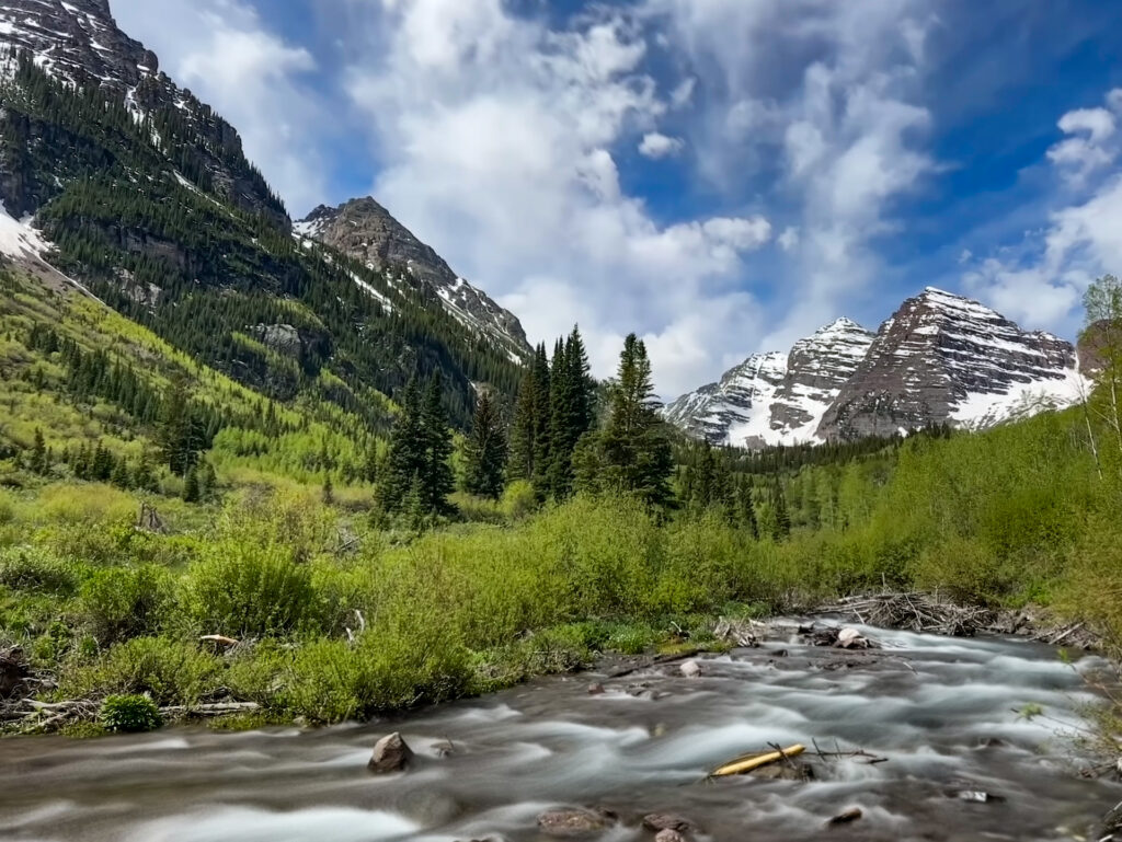 Maroon Bells Hike, Aspen, Colorado. A view of a stream and snow-capped mountains.